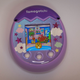 Photo of the purple Tamagotchi Pix laying on a table, with hearts surrounding the Tamgotchi character